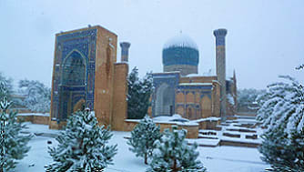 Historic Islamic architecture complex with blue dome and minarets surrounded by snow-covered evergreen trees in winter